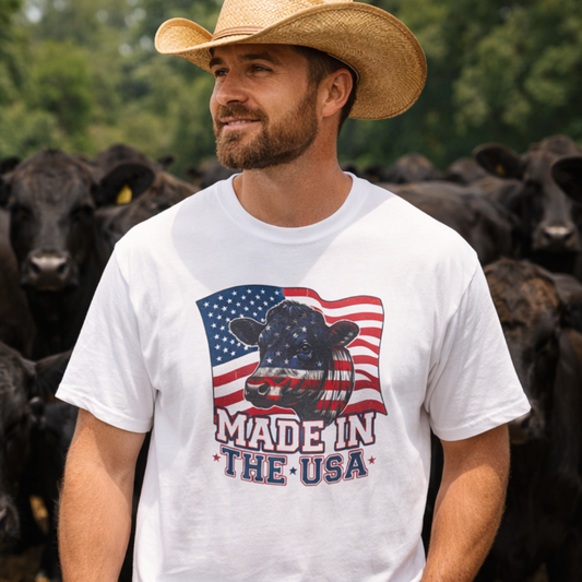 Man wearing a Rancher T-Shirt with a patriotic design, smiling outdoors among cattle.